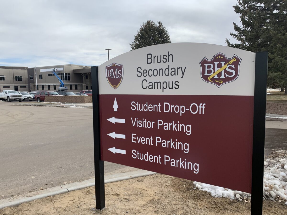 A directional sign at Brush Secondary Campus with arrows indicating locations for Student Drop-Off, Visitor Parking, Event Parking, and Student Parking. The campus building is visible in the background under a cloudy sky.