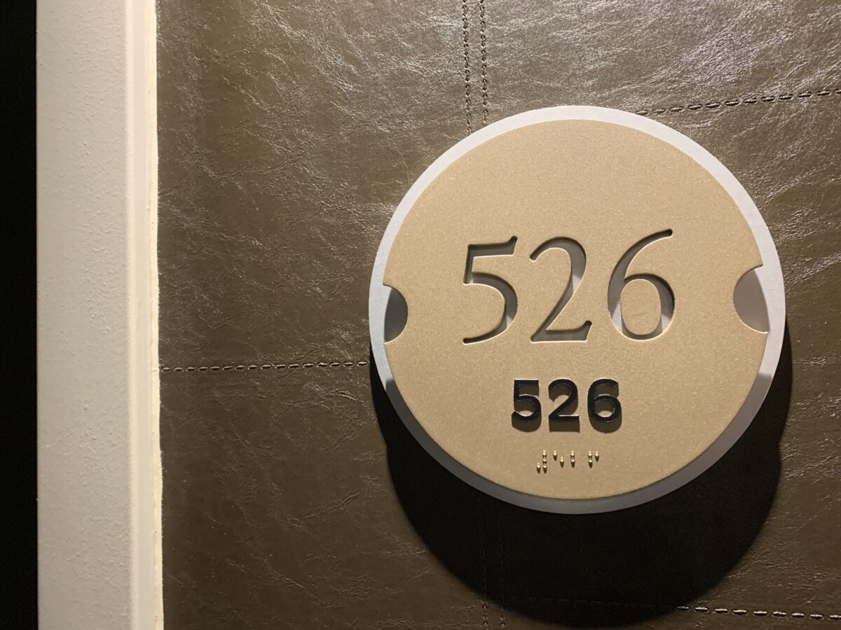 A round beige sign with the number "526" prominently displayed in black numerals is mounted on a gray-tiled wall. The number "526" appears again in smaller print below, with Braille dots below the smaller print.