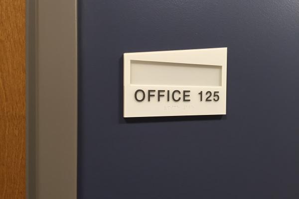 Minimalistic white sign mounted on a dark blue wall, labeled 'OFFICE 125' with a slot for a nameplate.