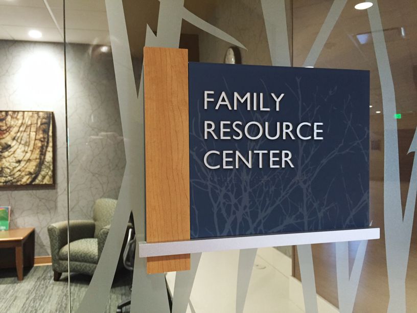 A glass door with a wooden and blue sign that reads "Family Resource Center." Inside, blurry furniture is visible, including an armchair and a wall painting. The background has a pattern of frosted stripes or branches on the glass.