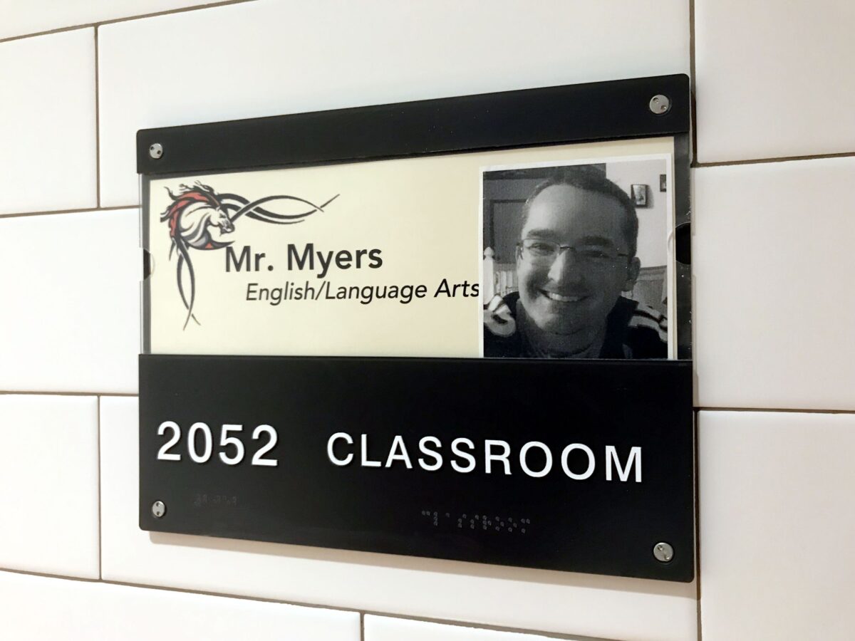 A classroom sign displays "2052 CLASSROOM" at the bottom. Above, there's a smaller sign that reads "Mr. Myers, English/Language Arts" with a profile picture of a man smiling. The sign is attached to a tiled wall.