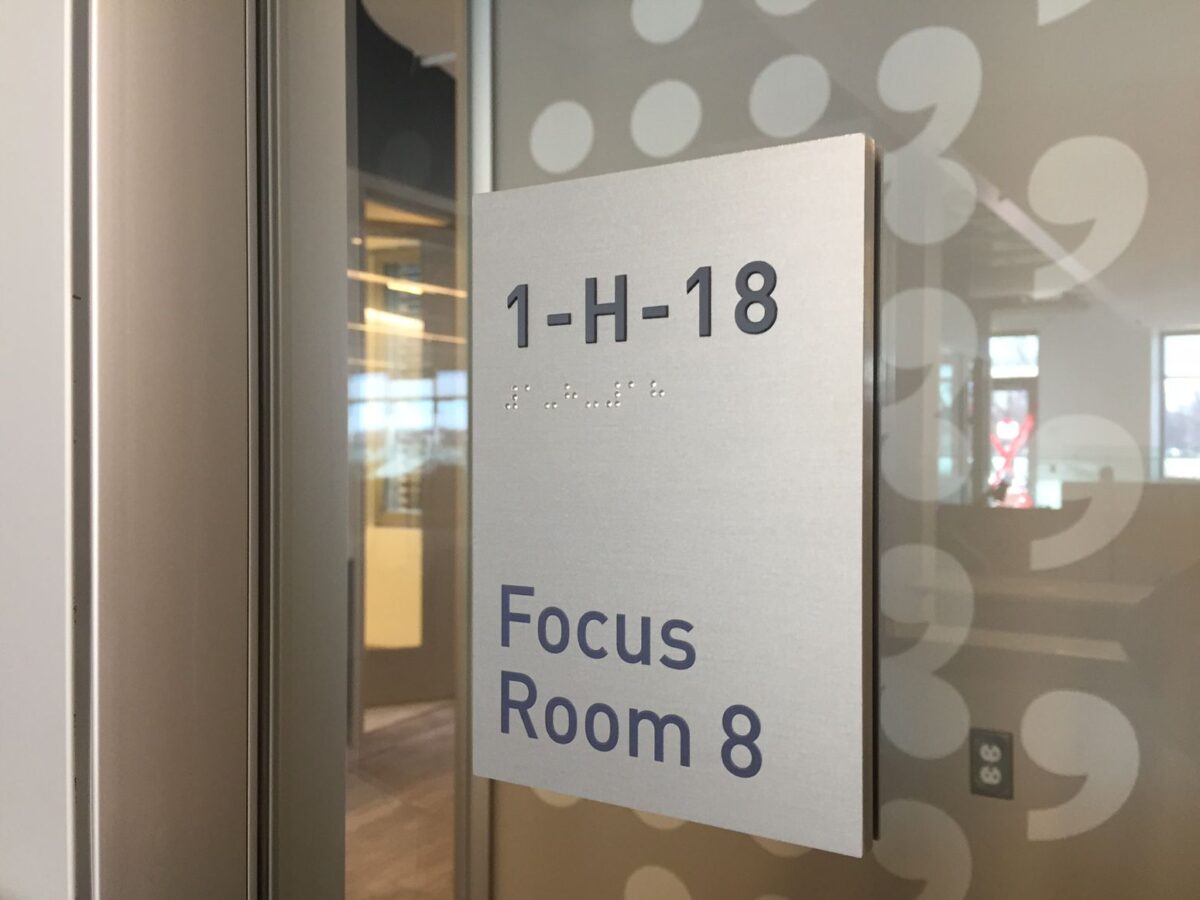 A sign on a glass door reads "1-H-18 Focus Room 8" in black text on a white background. The surrounding area appears to be an office space with a modern interior design, featuring large windows and bright lighting.
