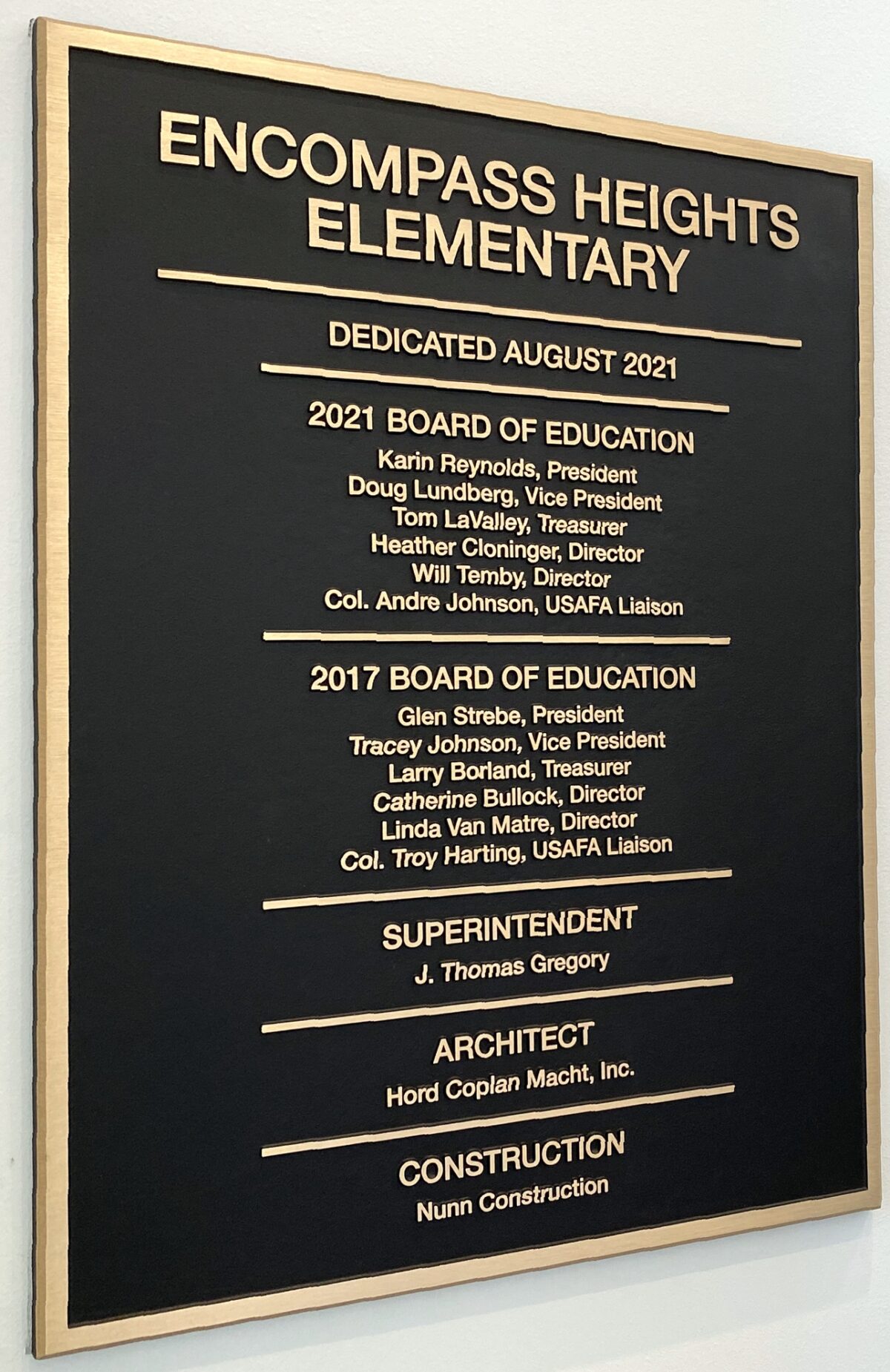 A plaque on a wall entitled "Encompass Heights Elementary," dedicated in August 2021. It lists members of the 2021 and 2017 Boards of Education, the Superintendent, and names the architect and construction company involved in the project.