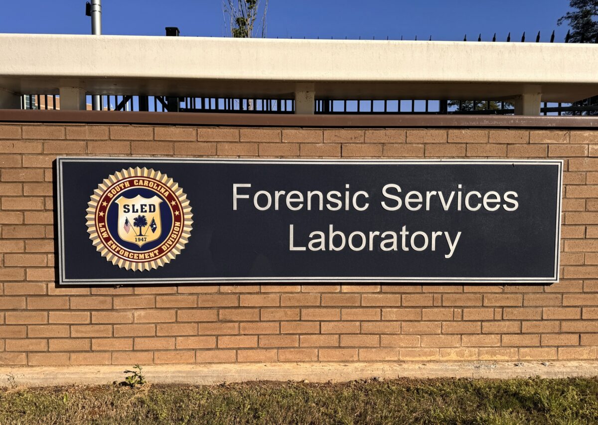 A brown brick wall features a large sign displaying the emblem and text that reads "SLED Forensic Services Laboratory." The sign is rectangular with a black background and white lettering. A patch of grass is visible in front of the wall.