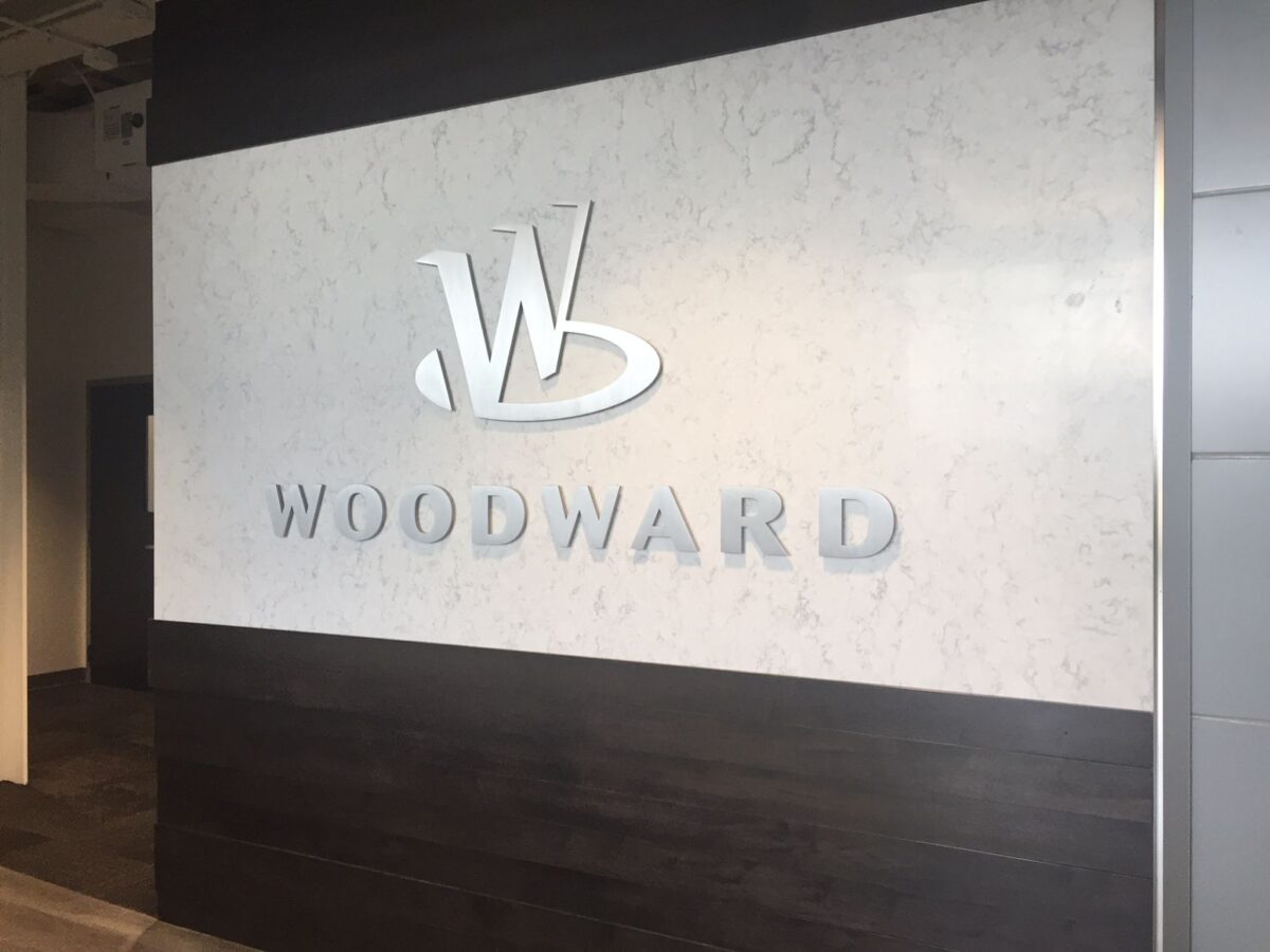 A wall with a sleek, modern design features the embossed logo and name of "Woodward." The logo consists of a stylized "W" above the name "Woodward." The background combines dark and light surfaces, adding contrast to the design.