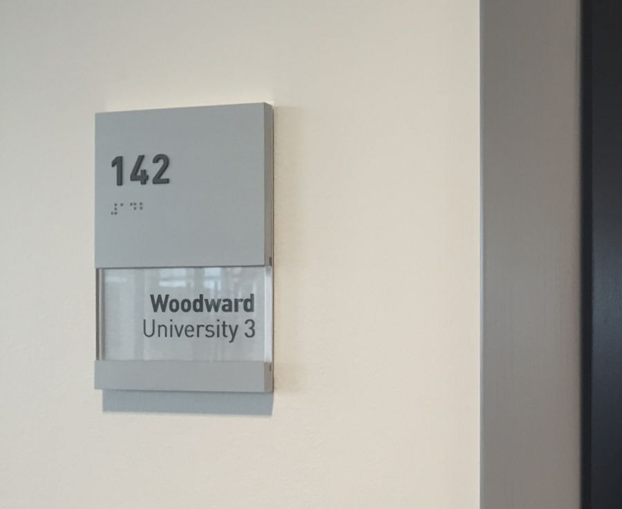 Room sign on a beige wall displays "142" in large numbers at the top left, "2nd Floor" in smaller text below it, and "Woodward University 3" in bold text on a clear panel beneath. The sign is gray with a slightly recessed text area.