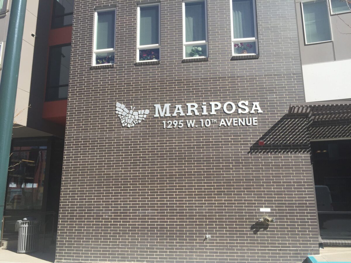 A brick wall with the text "MARIPOSA 1295 W. 10TH AVENUE" in white modern capital letters. To the left of the text is a white butterfly graphic. Shadows and sunlight create a pattern on the wall.