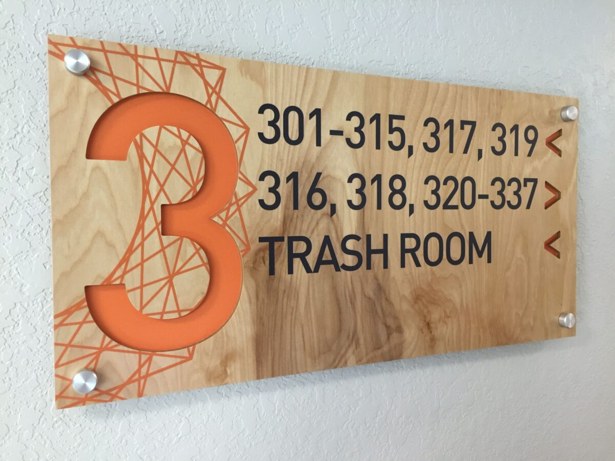 A wall-mounted wooden sign with an orange number 3 on the left, indicating the third floor. On the right, black text lists room numbers: 301-315, 317, 319, 316, 318, 320-337, and "TRASH ROOM" at the bottom. The sign has a decorative orange geometric design.