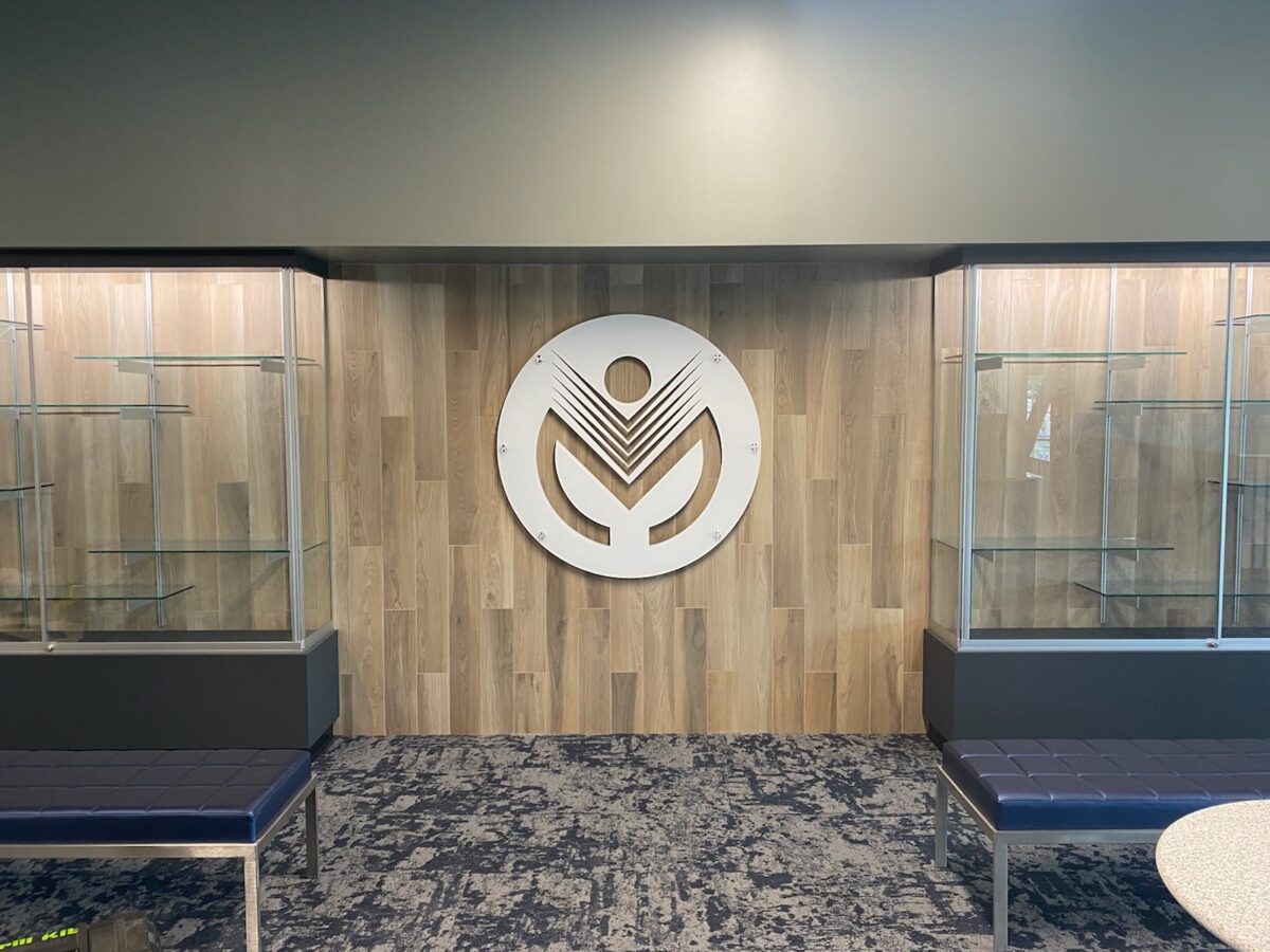 A wall with a wooden panel design displays a large, white, circular logo in the center. On either side of the logo are glass display cases with empty shelves. Blue cushioned benches with metal legs are positioned in front of the display cases.