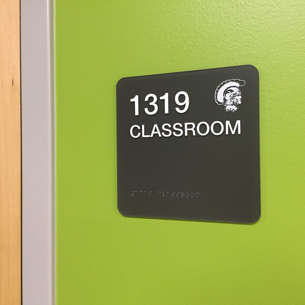 A green wall with a black sign displaying the room number "1319" in white text, above the word "CLASSROOM." The sign also includes a small image of a gladiator helmet and has Braille text at the bottom.