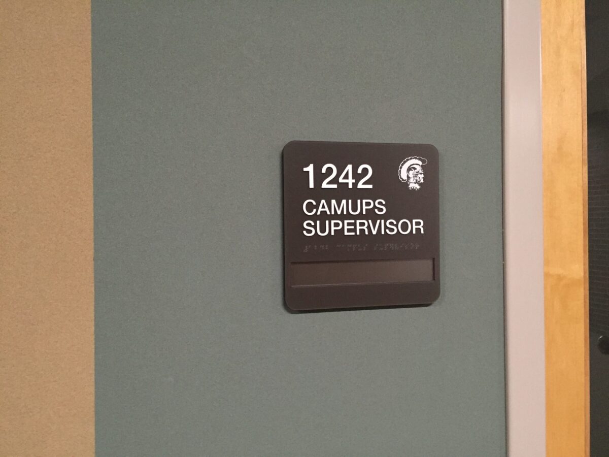 A wall-mounted black sign with white text that reads "1242 CAMUPS SUPERVISOR." There is also an icon of a head wearing a helmet above the text. The sign is mounted on a teal-colored wall.