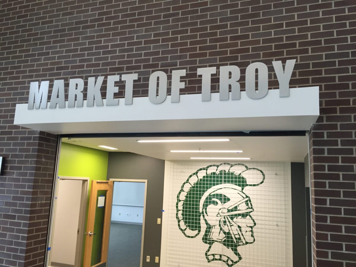 An entrance with a brick wall displaying the sign "Market of Troy" in large capital letters above it. Below the sign is a drawing of a Trojan helmet in green on a white tiled wall. To the left are open doorways leading into rooms painted in green and grey.