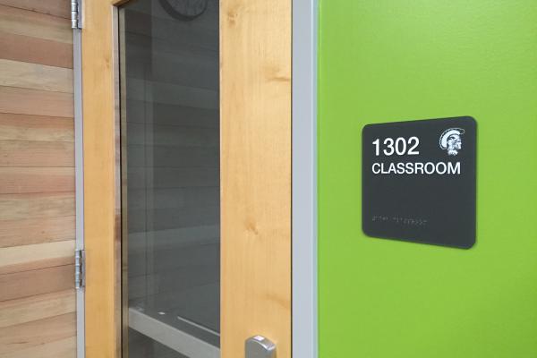 Modern classroom door sign labeled '1302 CLASSROOM' on a black background, mounted on a vibrant green wall next to a wooden and glass door.