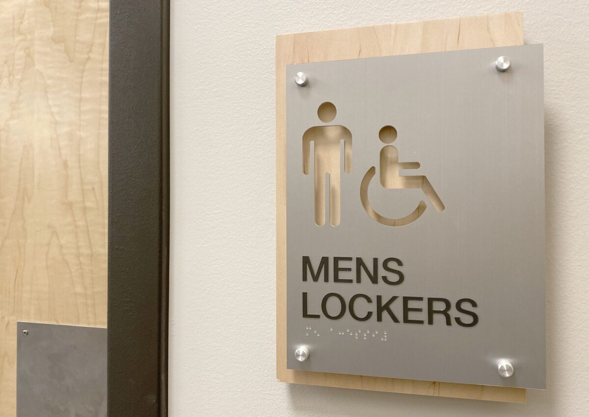 A sign on a wall indicates a men's locker room with accessible facilities. The sign features icons for a man and a person in a wheelchair above the text "Mens Lockers." The sign is mounted on a wooden panel next to a door.