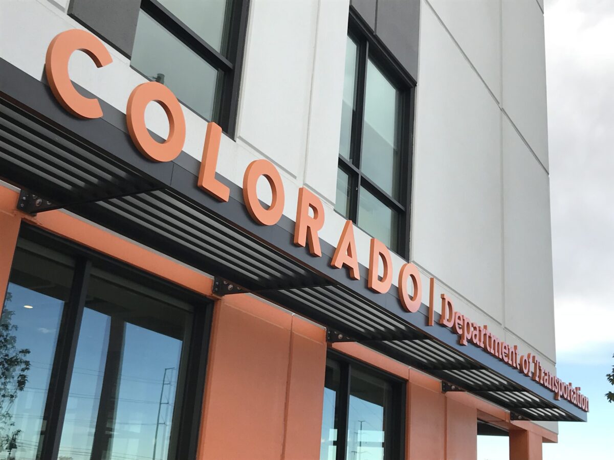 The image shows a building with a sign that reads "COLORADO Department of Revenue" in large, bold letters. The façade has a modern design with large windows and orange accents. The sky is partly cloudy in the background.