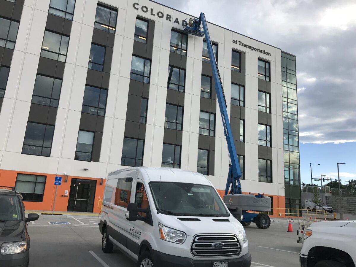 A white van is parked in front of a multi-story building with "Colorado" partially visible at the top. A blue construction lift is extended toward the building's higher floors. The building has large windows and a modern design. The sky is cloudy.