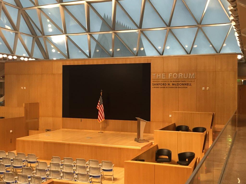 A modern, geometrically designed auditorium named "The Forum" with a large, blank screen flanked by wood paneling. An American flag stands beside a lectern on the stage. Rows of empty chairs and two smaller seating sections are visible.
