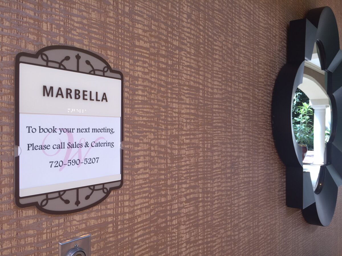 A sign on a patterned wall labeled "Marbella" with contact information for booking a meeting, instructing to call Sales & Catering at 720-904-5207. Next to the sign is a decorative, geometrically-shaped mirror reflecting some greenery.