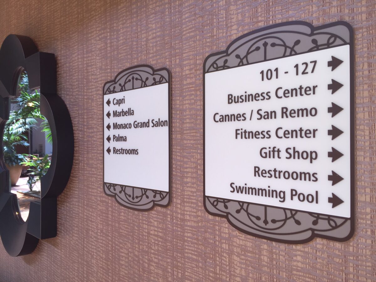 Two wall-mounted signage panels indicating directions in a facility. The left sign lists Capri, Murabella, Monaco Grand Salon, Primo, and Restrooms. The right sign points to rooms 101-127, Business Center, Cannes/San Remo, Fitness Center, Gift Shop, Restrooms, and Swimming Pool.