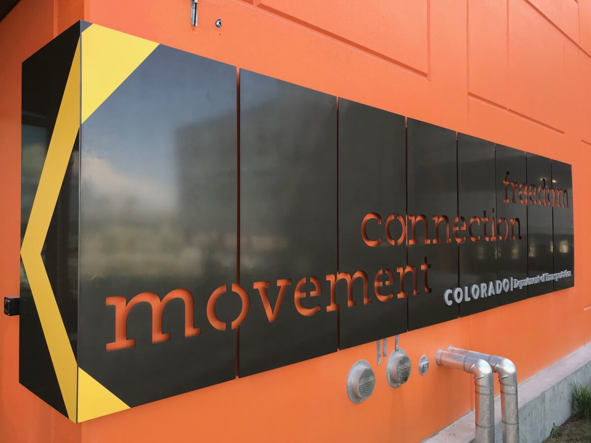 A large, rectangular black sign with yellow accents is affixed to an orange wall. The sign features the words "movement," "connection," and "Colorado" in orange and white text, partially obscured by reflections on the sign's surface.