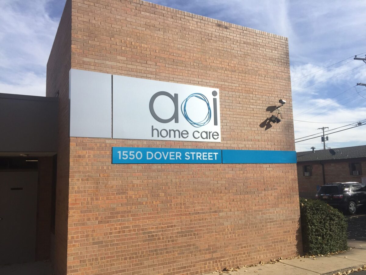 A brick building with a sign that reads "aoi home care" and a blue panel below it displaying the address "1550 Dover Street."
