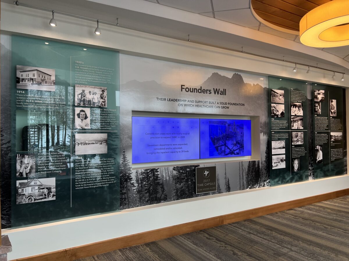 An indoor display titled "Founders Wall" with historical photos and text about the origins of a certain institution or place. The wall has a glass backdrop featuring a mountainous landscape and a central blue-lit screen with additional information.