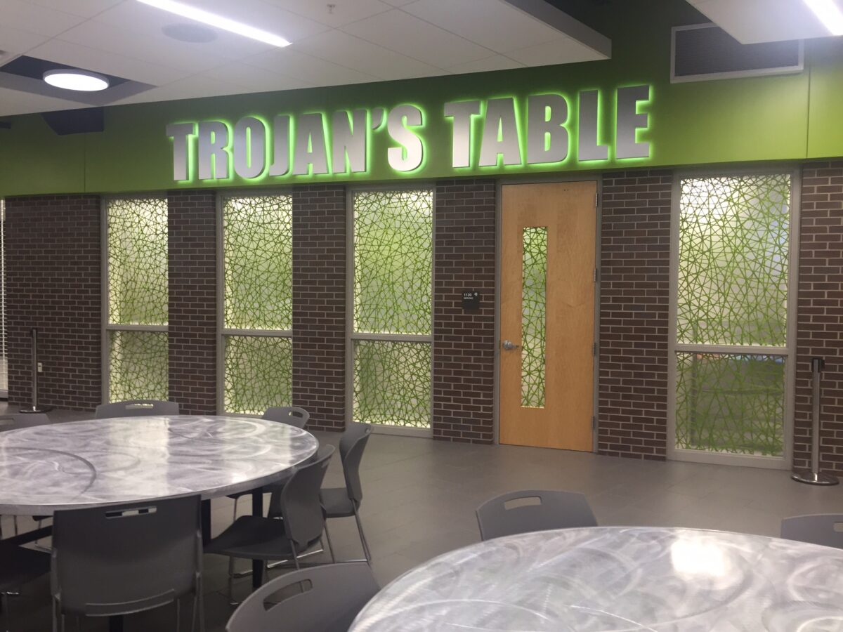 A dining area features a sign reading "TROJAN'S TABLE" in bold, green letters above frosted glass windows and a wooden door. The room includes round tables and chairs with a gray marble-like finish and modern design.