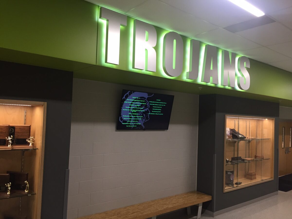 A hallway with a large illuminated sign reading "TROJANS" in green and white. Below the sign is a digital screen mounted on the wall. On either side of the hallway are display cases containing various trophies and awards. A wooden bench is positioned below the screen.