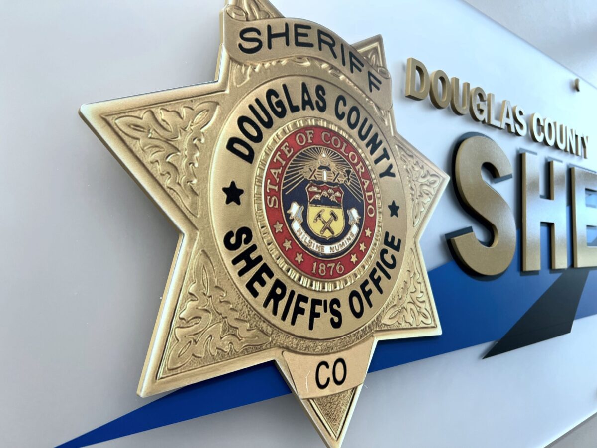 A gold badge with the text "Douglas County Sheriff's Office" and "State of Colorado" is mounted on a wall. The wall also displays large, metallic letters spelling "Sheriff" beside the badge. The badge features intricate design details and the year "1876.