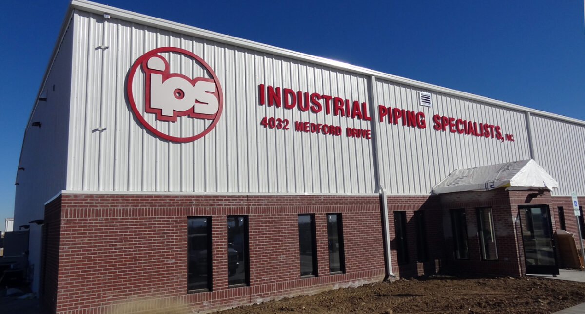 A large industrial building with "IPS" logo and text reading "Industrial Piping Specialist, Inc." on the white upper facade above red brick lower walls. The building is located at "4025 Menoah Road" with blue sky in the background.