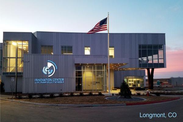 Dusk view of the Innovation Center by St. Vrain Valley Schools in Longmont, CO, featuring a modern, metallic building with a large logo, glass facade, and an American flag waving at the front.