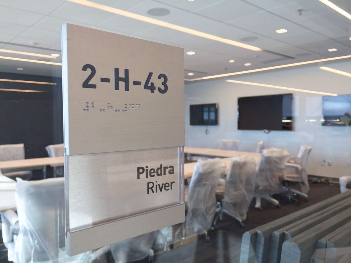 A modern conference room with plastic-covered chairs surrounding a table. The room's glass door has a sign displaying "2-H-43" with Braille text below and "Piedra River" written underneath. Several screens are mounted on the walls inside.