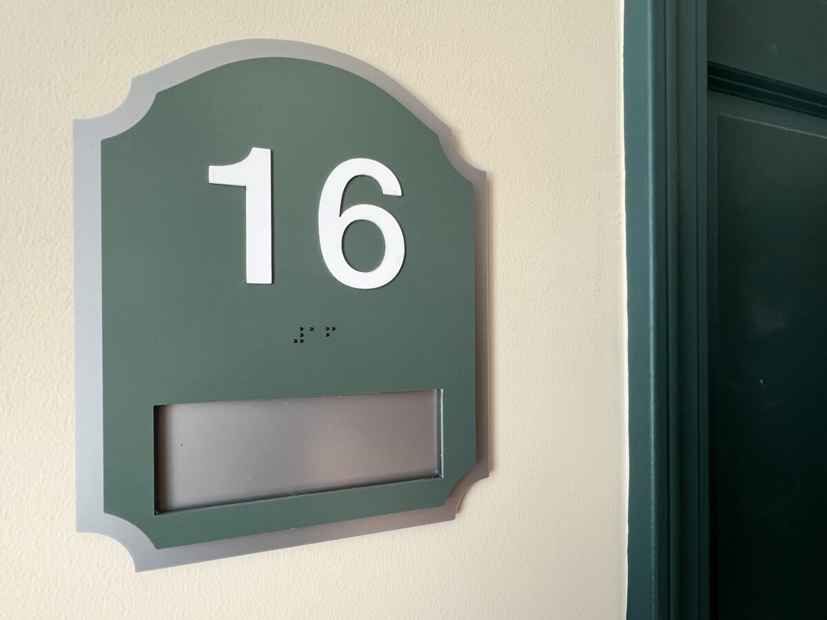 A green and gray door sign with the number 16 prominently displayed in white. Below the number, there is a small line of braille dots. There is a blank rectangular slot near the bottom of the sign. The door next to the sign is slightly ajar.