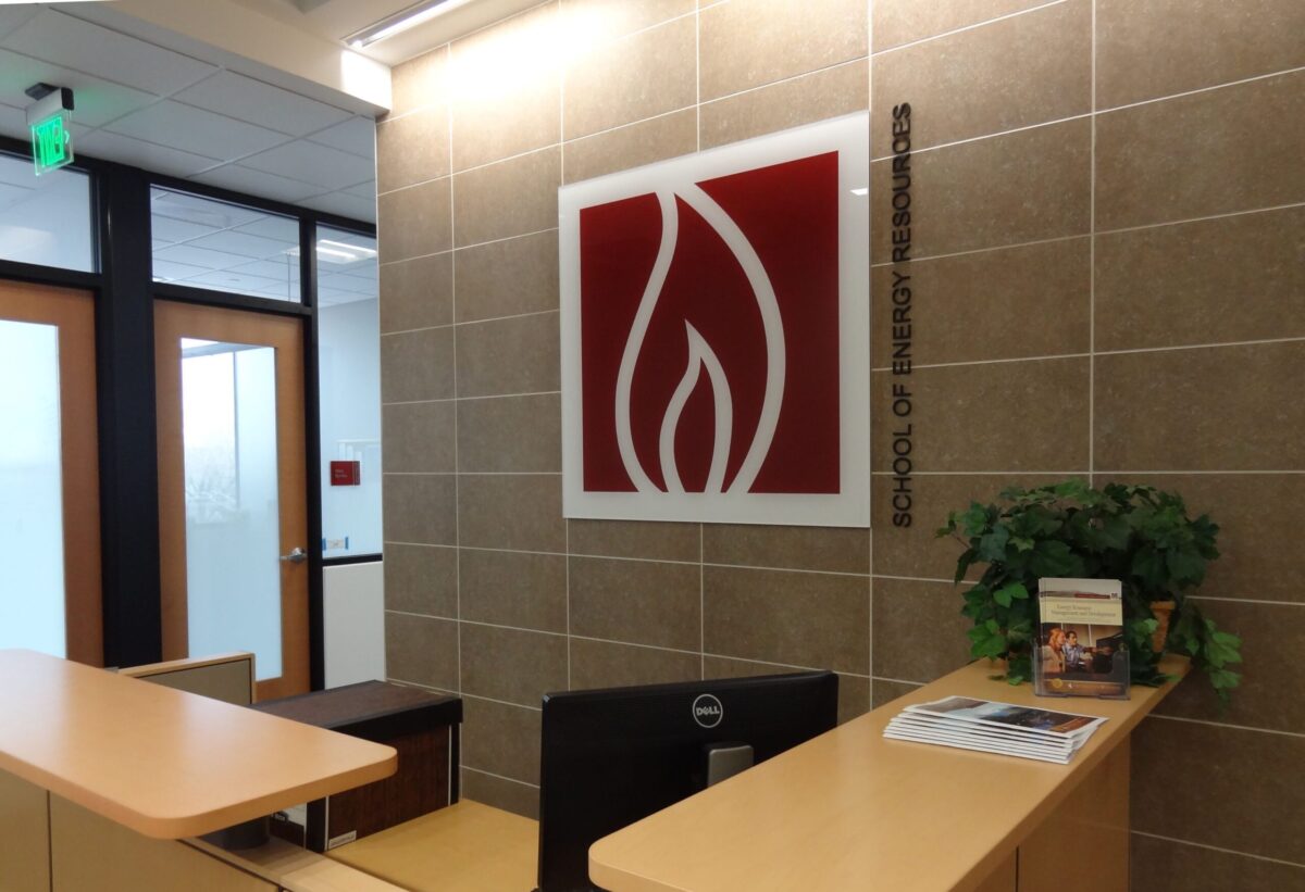 A modern office reception area with a wooden desk, computer screen, and a plant. Behind the desk on the wall is a large red and white logo featuring a stylized flame. Frosted glass doors and a green exit sign are visible in the background.