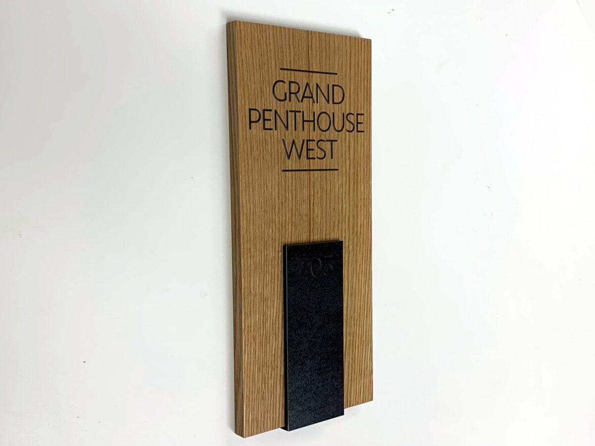A wooden sign for "Grand Penthouse West" mounted on a white wall, featuring a minimalistic design with black text and a small black vertical strip at the bottom.