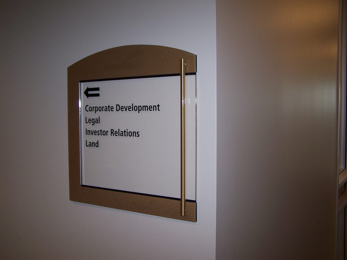 A directory sign on a wall points left with an arrow and lists the departments: Corporate Development, Legal, Investor Relations, and Land. The sign is mounted within a brown wooden frame.