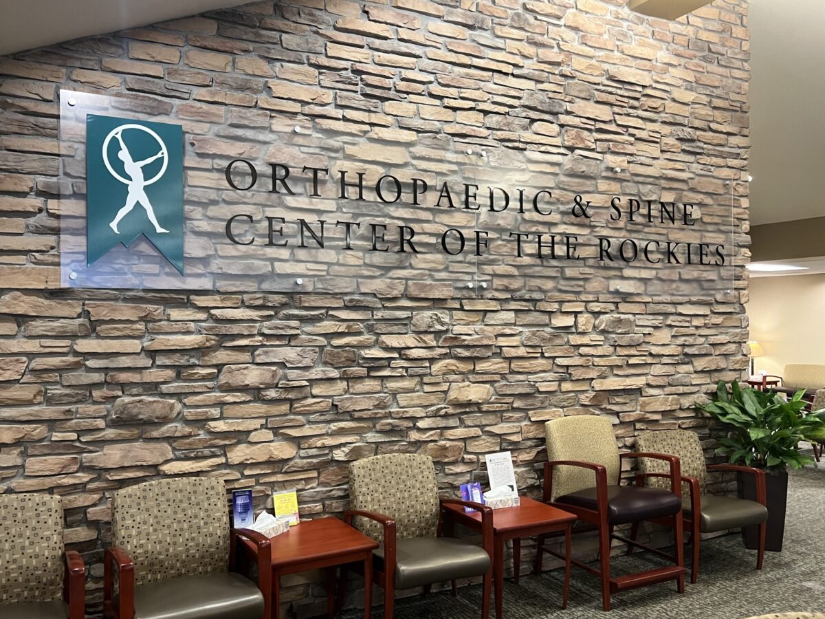 Lobby of the Orthopaedic & Spine Center of the Rockies, featuring a stone wall with the center's logo and name, and a seating area with several chairs and small tables.