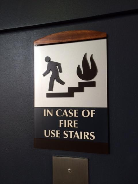 Safety sign 'In Case of Fire Use Stairs' with graphical representation of a person descending stairs next to a fire symbol, on a dark background with wooden framing.