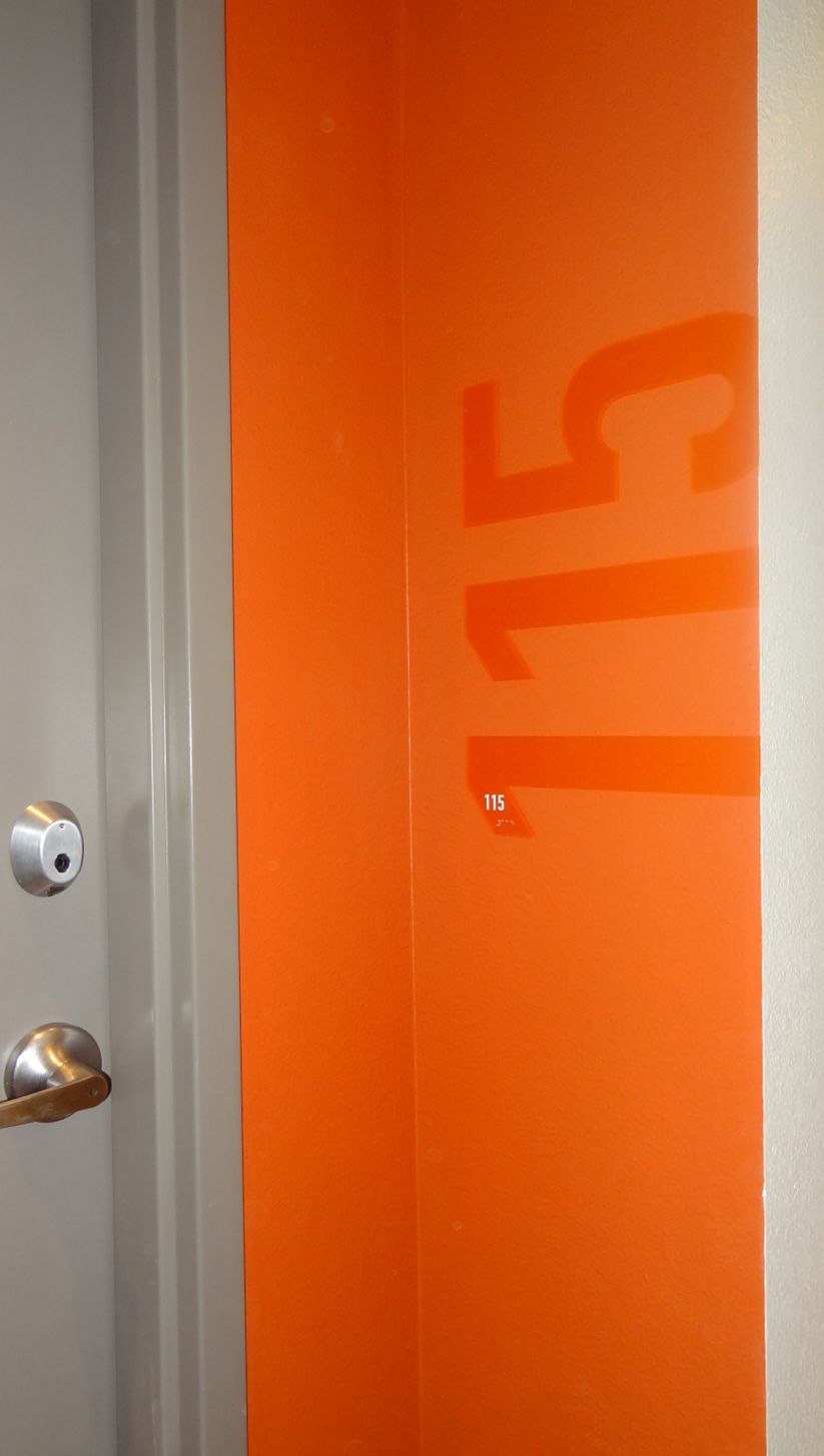 A door with a silver handle and lock is partially open, revealing an orange wall inside with the number "715" written vertically in large, glossy numbers. The door frame is grey.