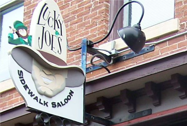A sign for "Lucky Joe's Sidewalk Saloon" is mounted on a brick building. The sign features a caricature of a cowboy wearing a hat. A streetlamp with a curved arm is attached to the same structure above the sign.