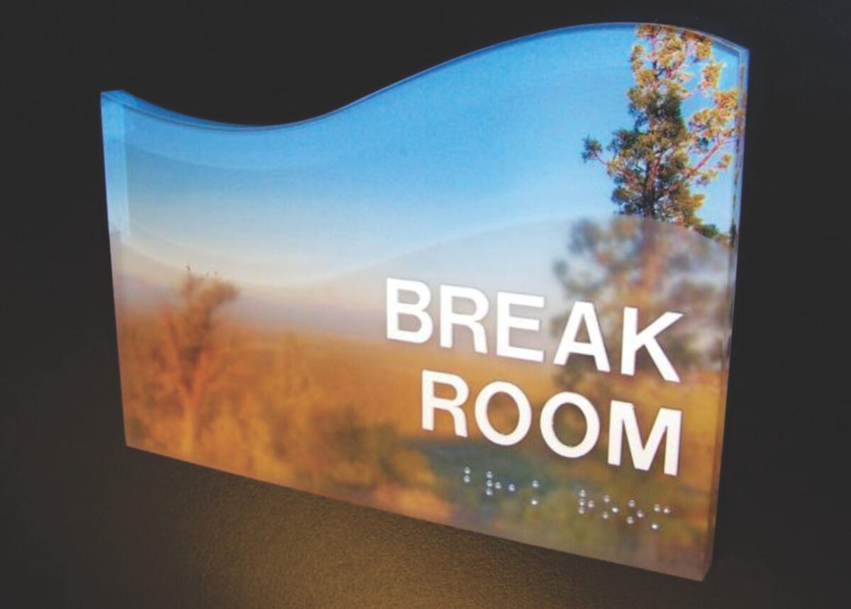 A sign with a scenic background featuring trees and a sky. The sign reads "BREAK ROOM" in large, white letters, and there is Braille text beneath the words. The top edge of the sign has a wave-like curve.