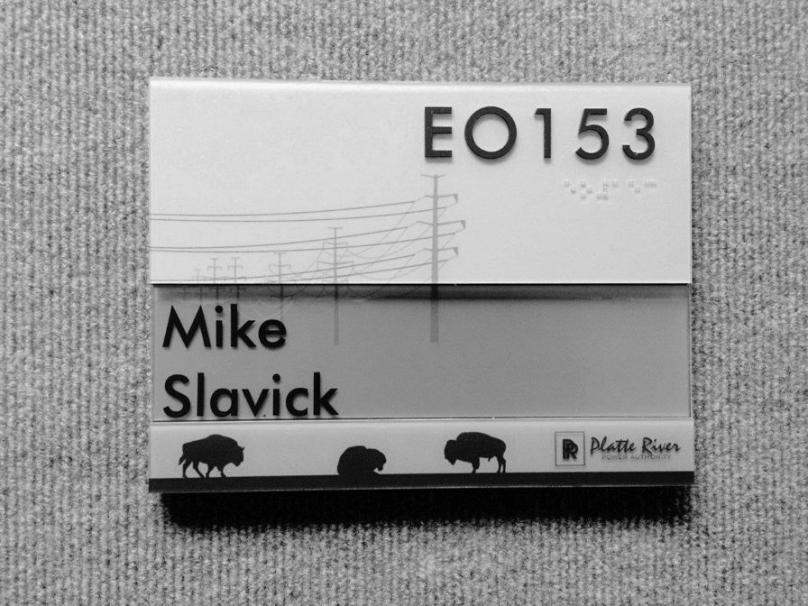 Office nameplate on a gray fabric background, featuring the text "EO153" and "Mike Slavick." The design includes illustrations of power lines and three buffalo silhouettes at the bottom. The Platte River logo is also visible.