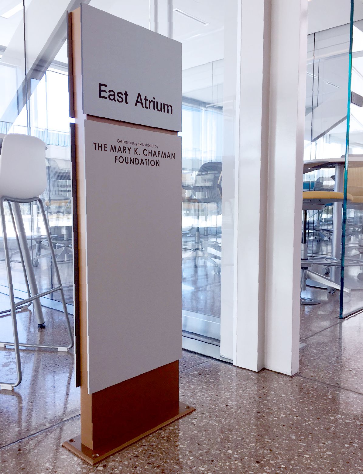 A sign in a modern indoor space reads "East Atrium" and "The Mary E. Chapman Foundation." The background shows a glass doorway with reflections and chairs along the left side. The polished floor reflects parts of the surroundings.