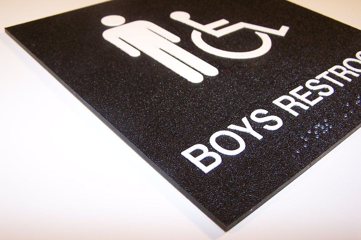 A black, textured sign with white icons indicating a boys' restroom. The sign includes a male figure and a wheelchair symbol, with the text "BOYS RESTROOM" written below. The sign is slightly angled, showing the texture clearly.