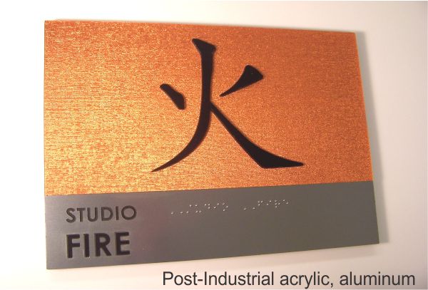 A sign with a metallic orange background features a large black kanji character for "fire." Below, on a silver and gray section, the text reads "STUDIO FIRE" in black capital letters. There is also a section with Braille text below the written text.
