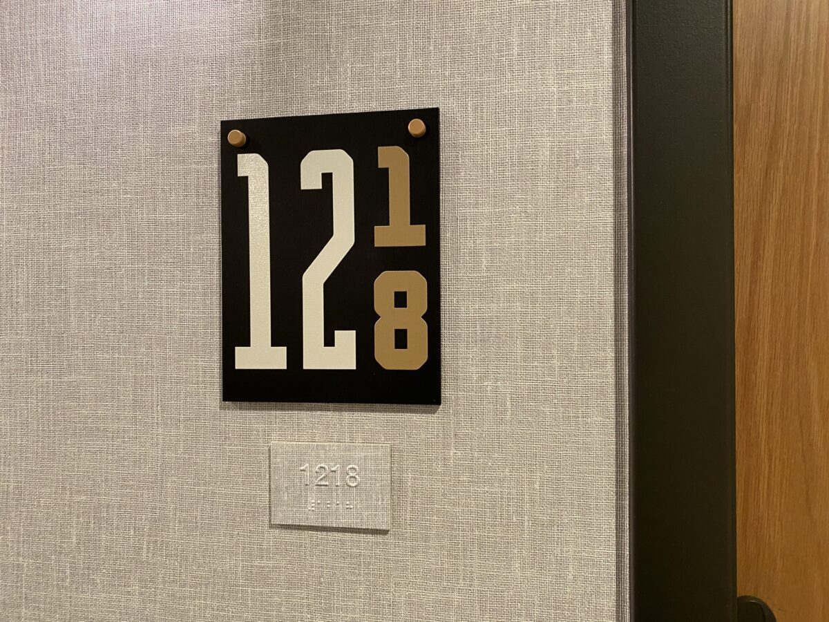 A wall-mounted room number sign reads "1218," with a unique design where the number "1" is smaller and raised above the "2" in the sequence. Below the sign, there's a smaller rectangular plaque displaying "1218" in Braille. The wall has a textured pattern.