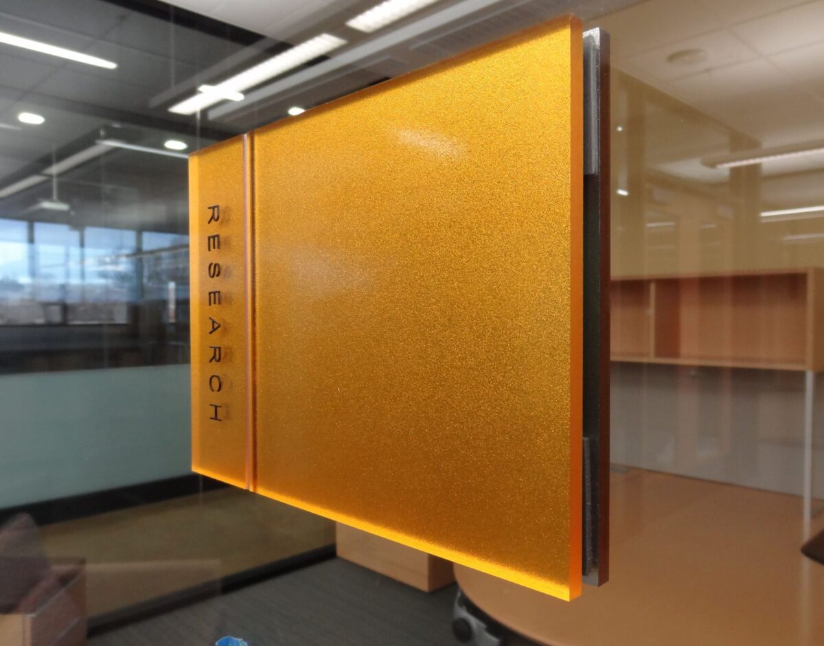 Frosted glass wall sign with orange tint and rounded corners, featuring the word RESEARCH in vertical text in black. The background shows a modern office interior with glass walls, a round table, chairs, and overhead fluorescent lighting.