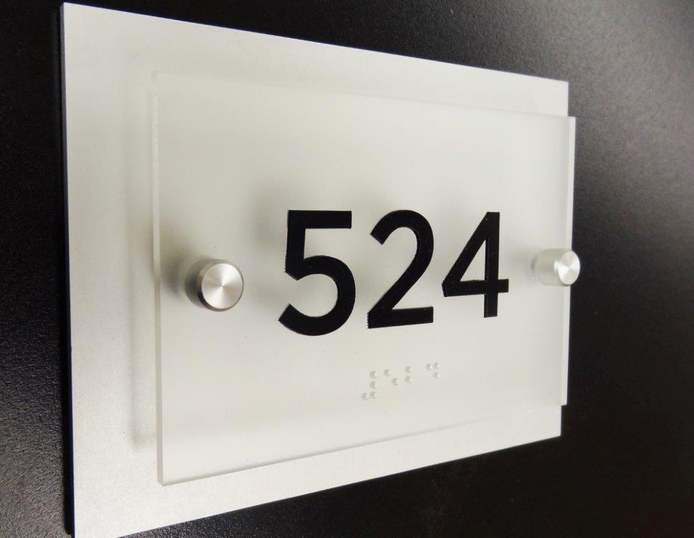 A room number plaque against a black background displays the number "524" in large black font. The plaque is made of frosted glass with two metal bolts securing it to the wall, and includes Braille text below the number.