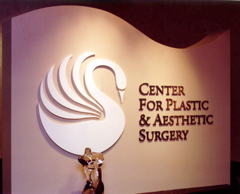 Sign for the "Center for Plastic & Aesthetic Surgery" displayed on a wall. The sign features a stylized white swan logo on the left and the center's name in dark letters on the right. An abstract metallic sculpture is placed on a shelf below the sign.