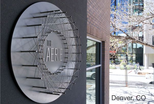 A circular metal sign displaying "The Aerie" is mounted on the side of a building. The sign features a radial design with a geometric pattern. In the background, there are urban buildings and a partial view of tree branches. Text at the bottom reads, "Denver, CO.